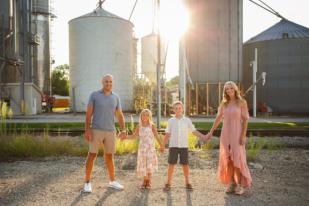 Family-of-four-holding-hands-and-smiling-in-front-of-industrial-silos-during-a-rustic-outdoor-photoshoot-in-soutside—indianapolis-bargersville