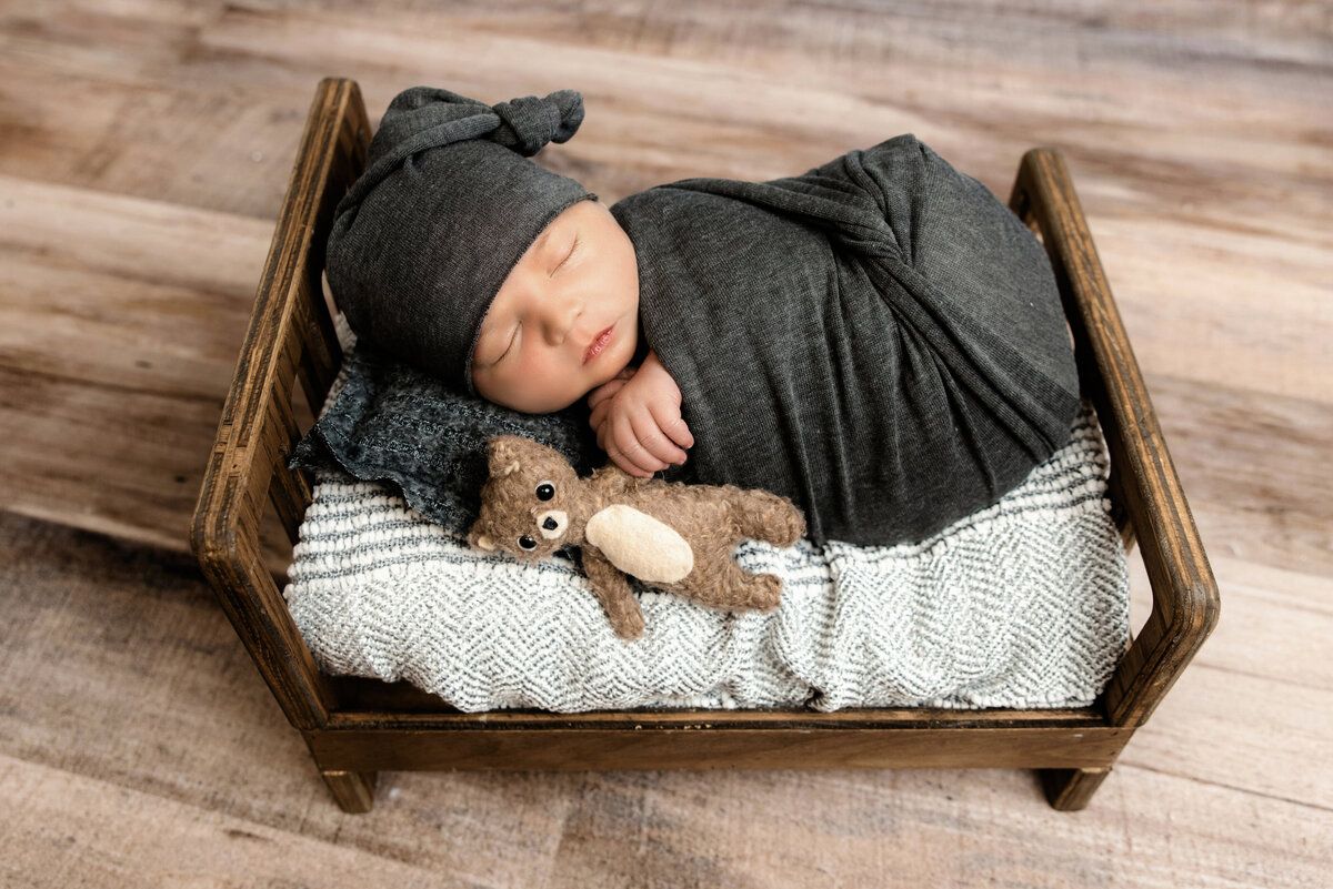 baby boy wrapped  in charcoal gray with sleepy and stuffed teddy on tiny brown bed