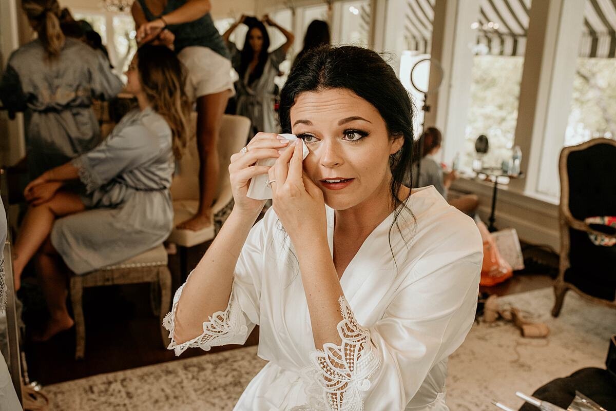 Bride touching up makeup after an emotional moment while sitting in the bridal suite wearing a white silk robe with lace details at Church of the Assumption in Nashville, TN