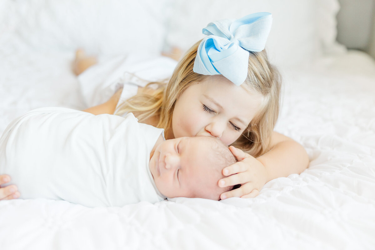 big sister kissing newborn baby boy on bed in swaddle in Goldsboro, NC