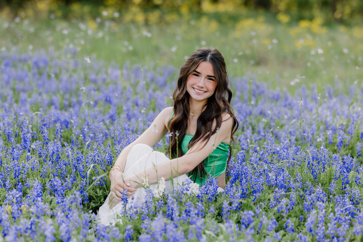 portrait of girl in green top and white pants sitting in field of bluebonnets