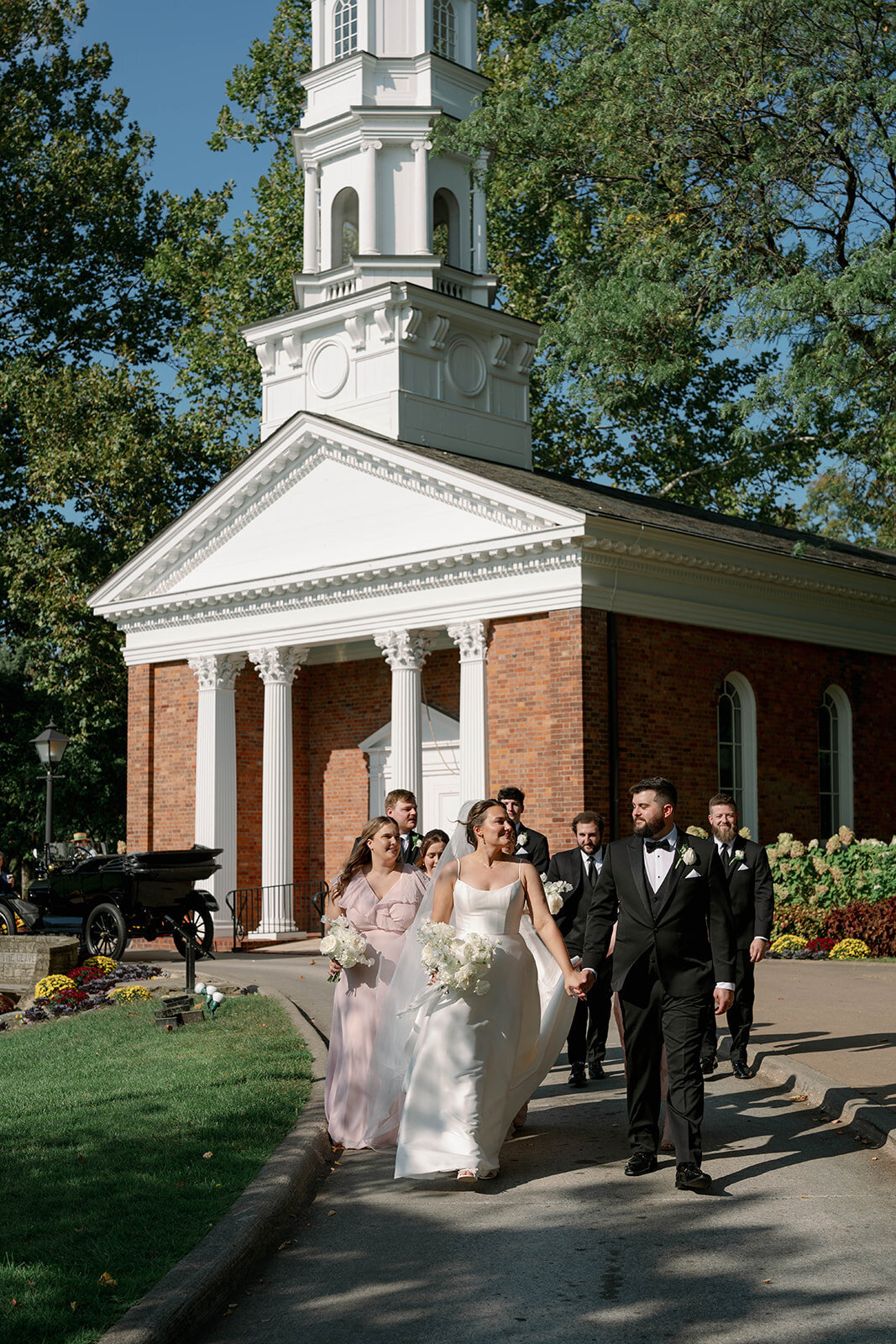 Bride walking with bridesmaids in soft pink dresses at Dearborn Village after Martha-Mary Chapel ceremony, candid wedding party photo.