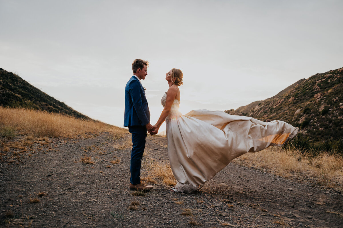 Bride and groom stand facing each other while her satin gown blows in the wind in front of sunset.