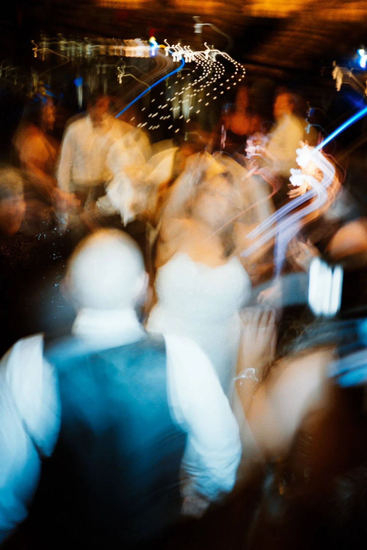 A blurred, abstract photo of people dancing at a lively event, captured by a film photographer NJ, with colorful light trails and indistinct figures creating a sense of motion and energy.