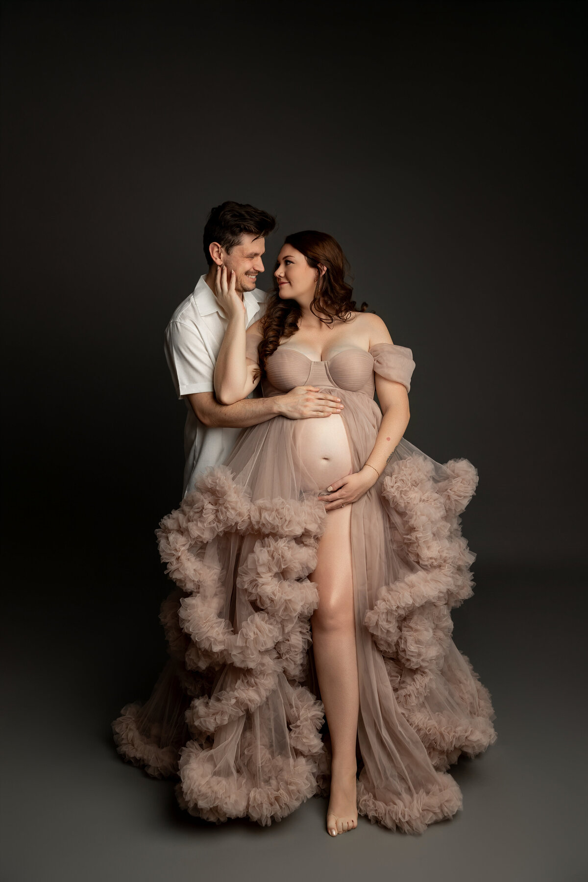 The expectant mother, wearing a soft beige dress, lovingly touches her husband's face, creating a tender and intimate moment between the couple. Captured by Inland Empire maternity photographer Kaitlyn Dawn Photography.