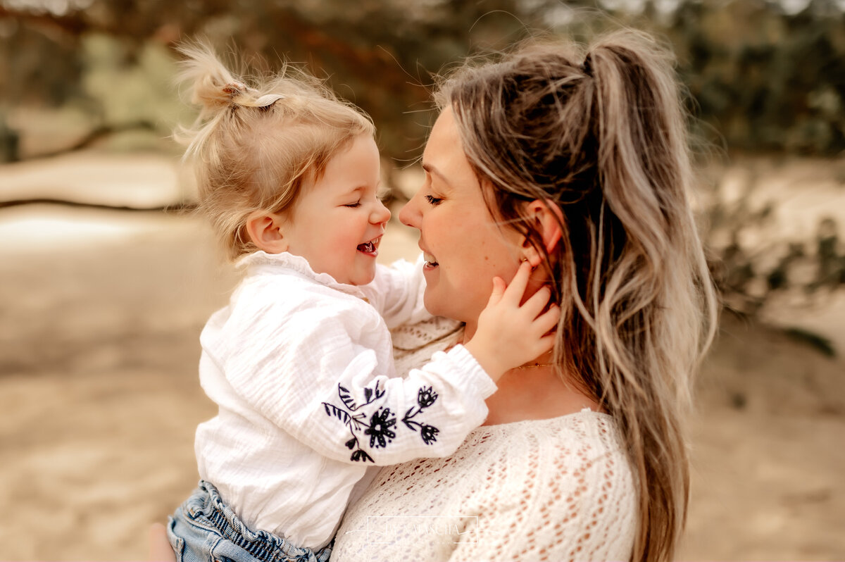 Mama en dochter knuffelen met neusje tegen neusje en lachen naar elkaar in duinen Herperduin
