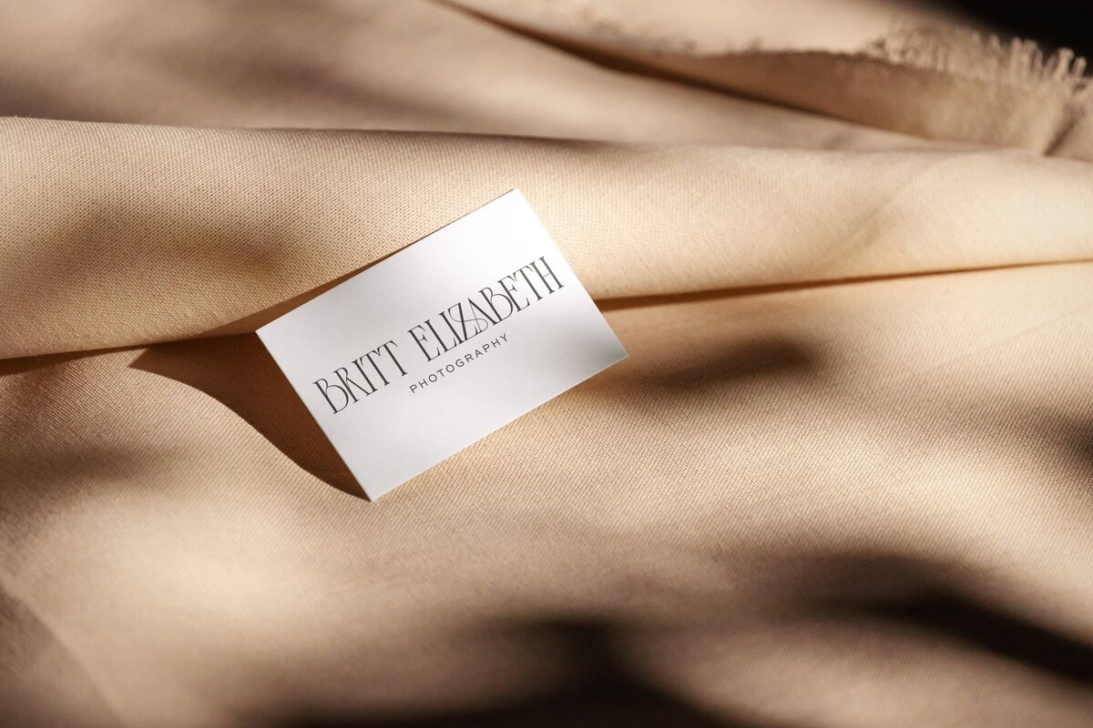 White business card with "Britt Elizabeth Photography" text placed on a draped beige fabric under soft lighting, showcasing elegant simplicity that aligns perfectly with our branding and website design for photographers.