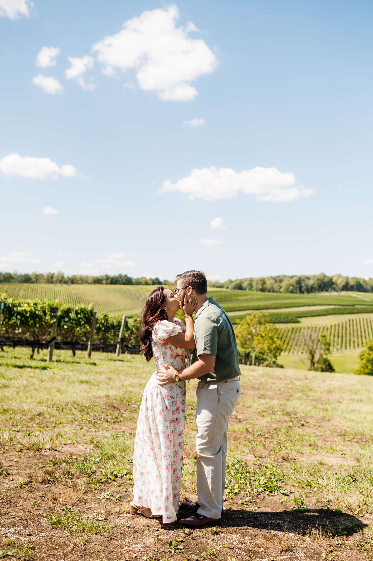 white dress for engagement photos on a brunette woman who is holding her fiances face and kissing him with a vineyard and rolling hills behind them as the man holds to woman's waist