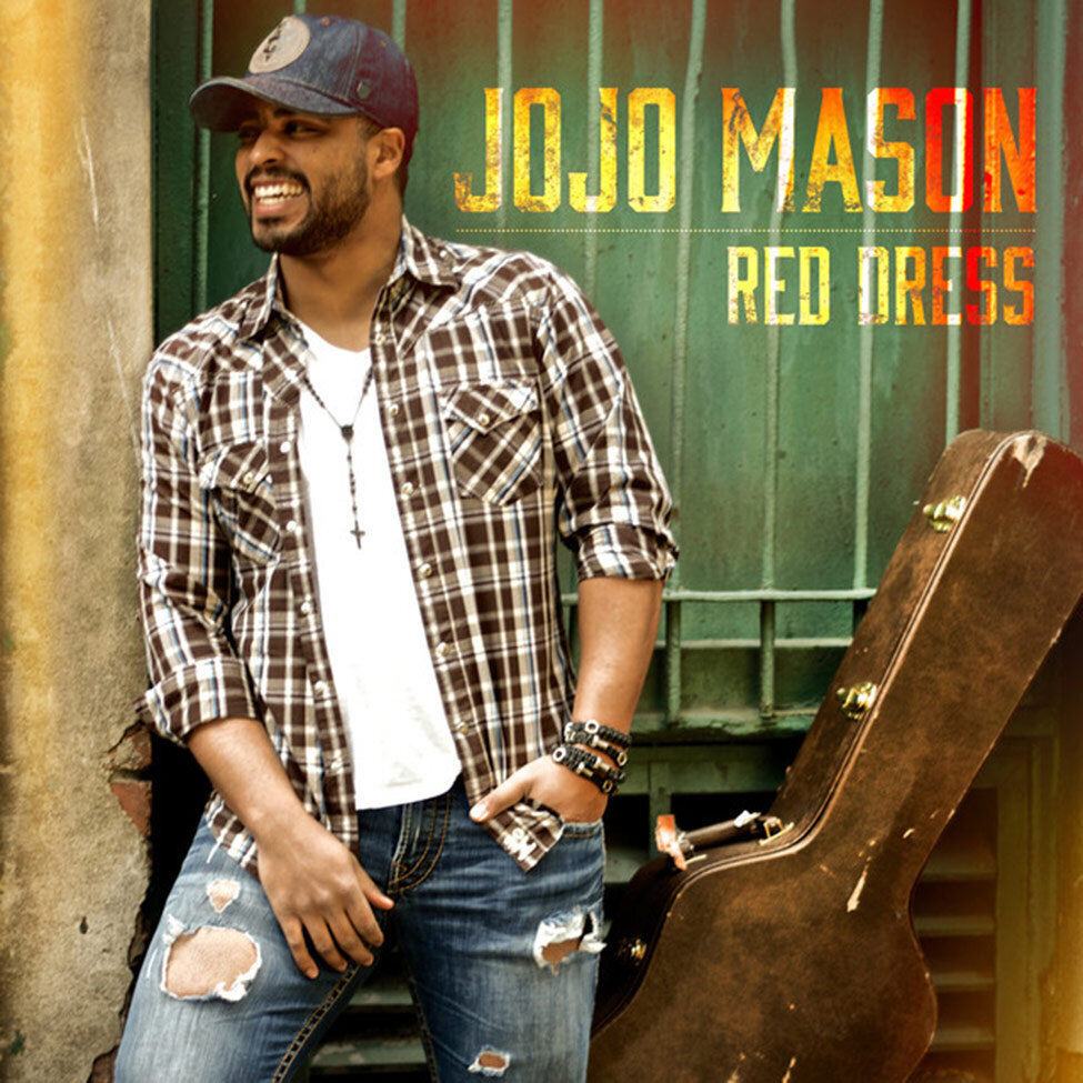Single Cover for Red Dress by JoJo Mason  singer smiling while standing against brick wall and green window with bars one hand in pocket guitar case leaning on wall beside him