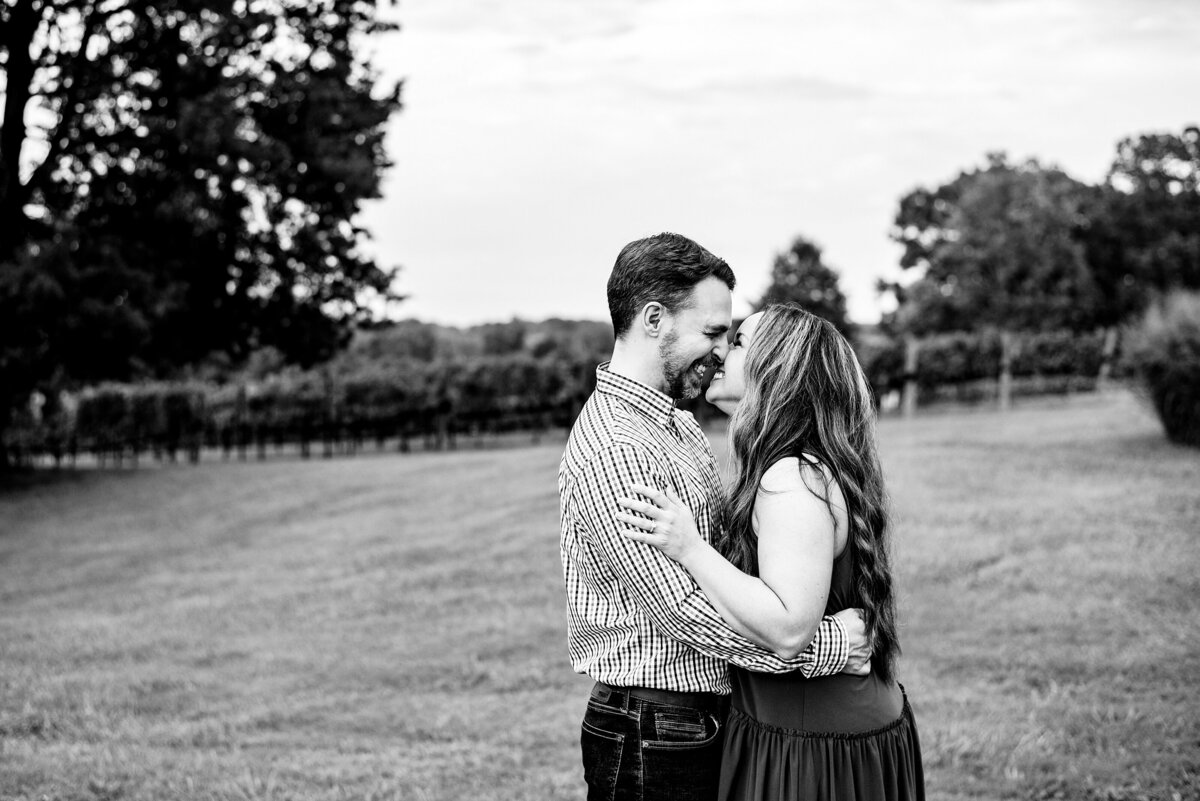 Black and White photo of couple nuzzled together smiling and about to share a kiss with the hills of Arrington Vineyard behind them