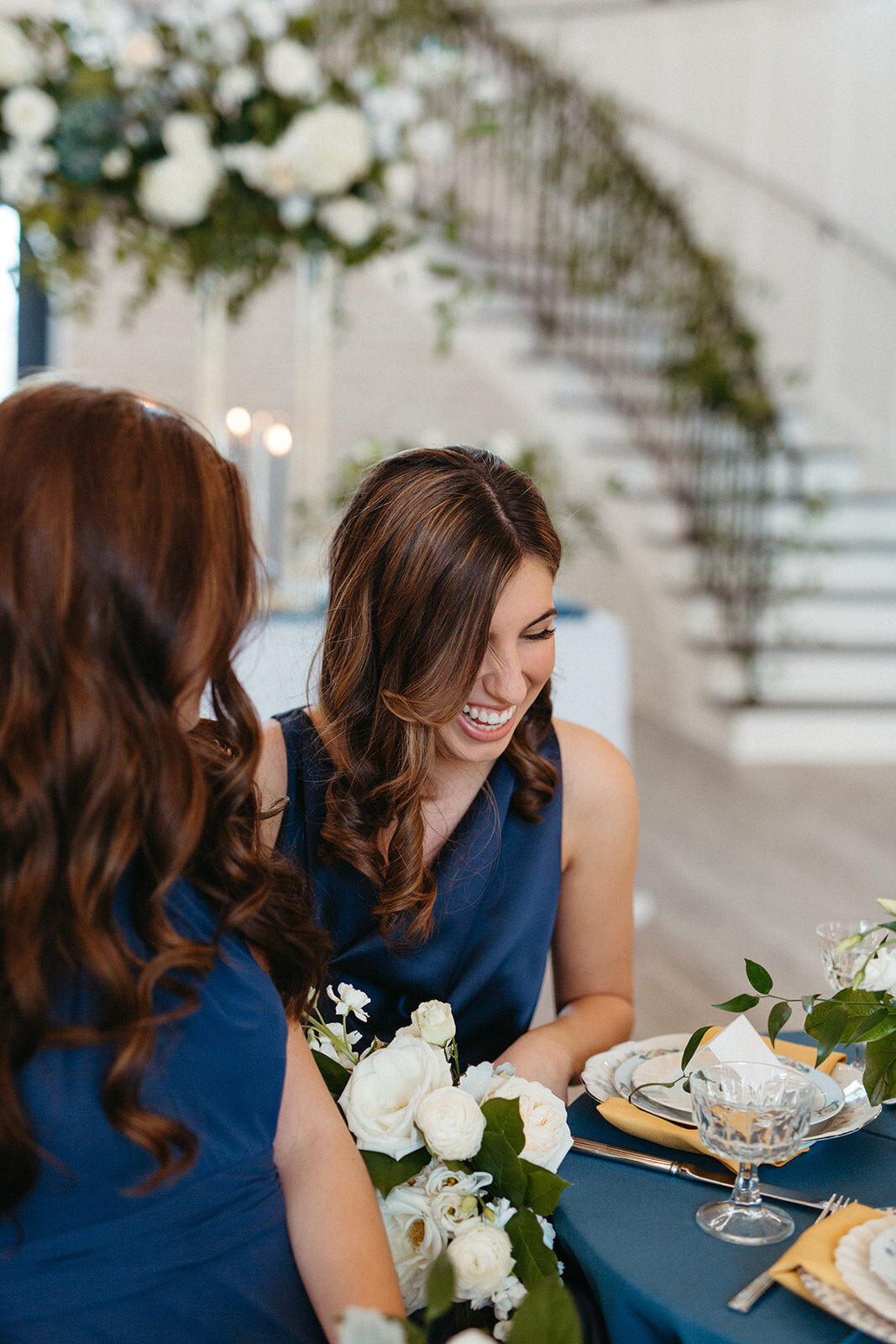 Two bridesmaids wearing blue satin gowns smile while seated at a table in the banquet room.