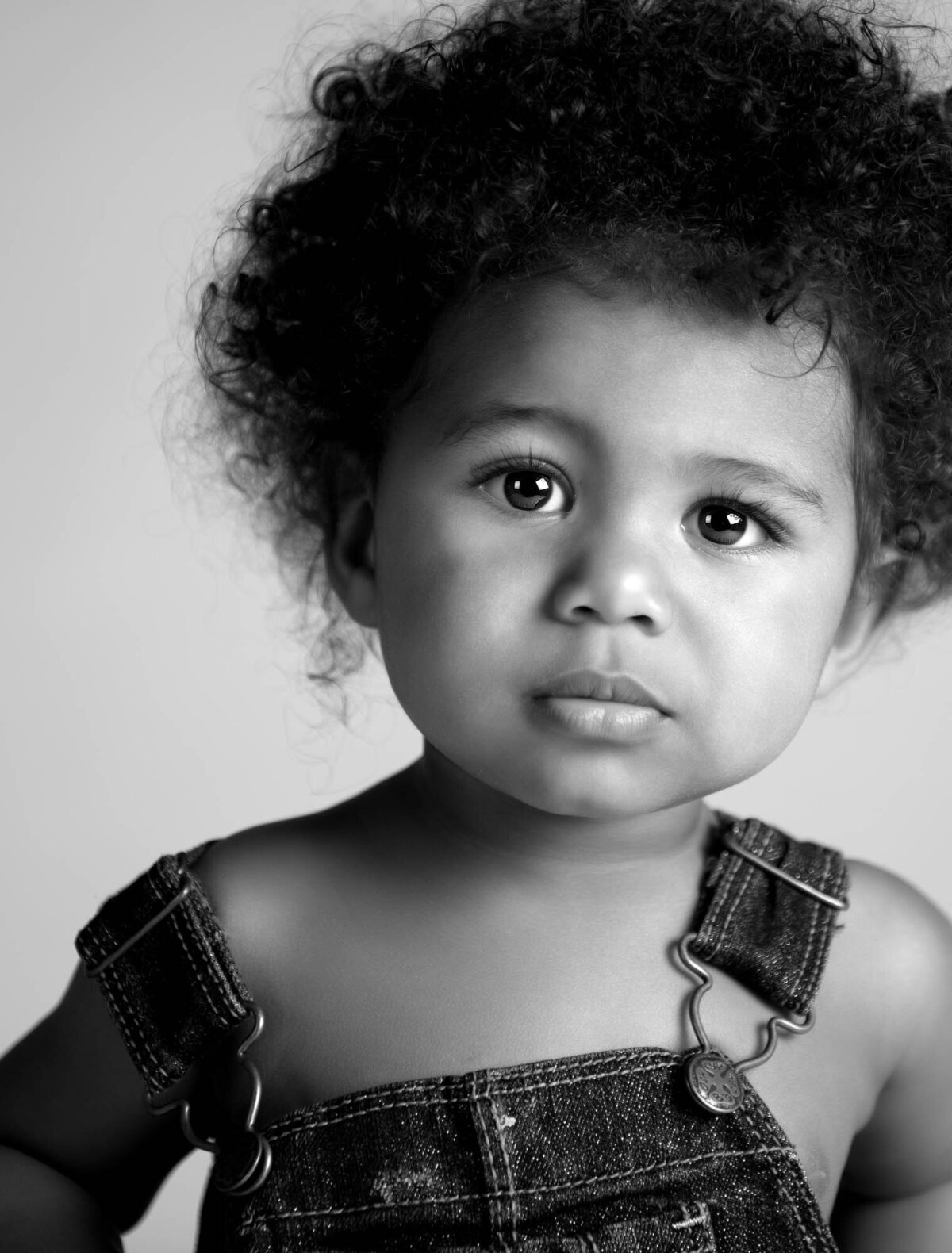 A black-and-white portrait of a young child with curly hair and a serious expression. The child is wearing denim overalls and looking directly at the camera.