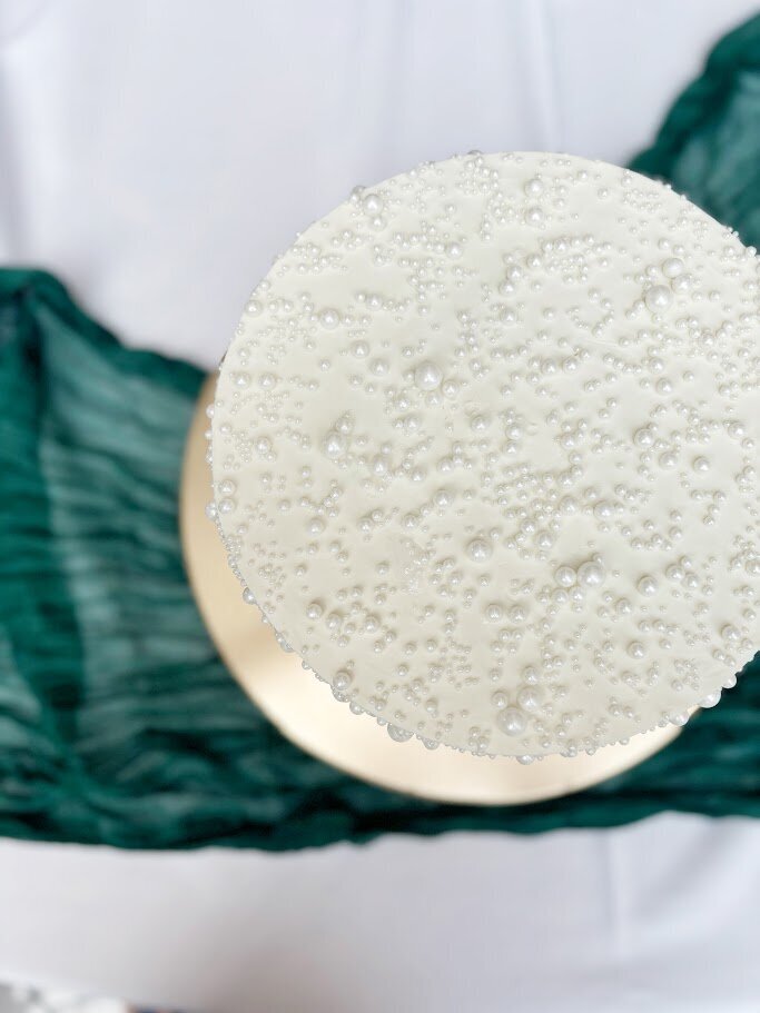 A shot of the top of a wedding cake, overed with different sizes of sugar pearls. An emerald green cloth is seen underneath.