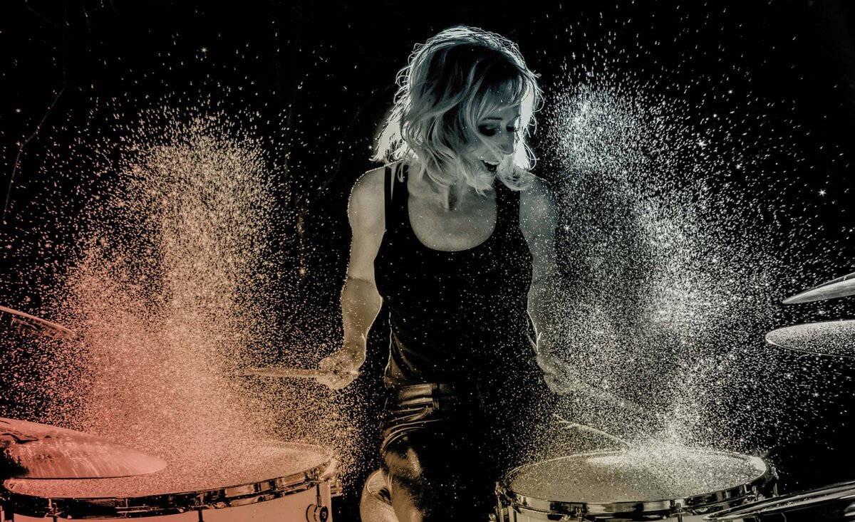 Music portrait Andra Liemandt playing drums wearing black tank top golden pink dust flying up from drums