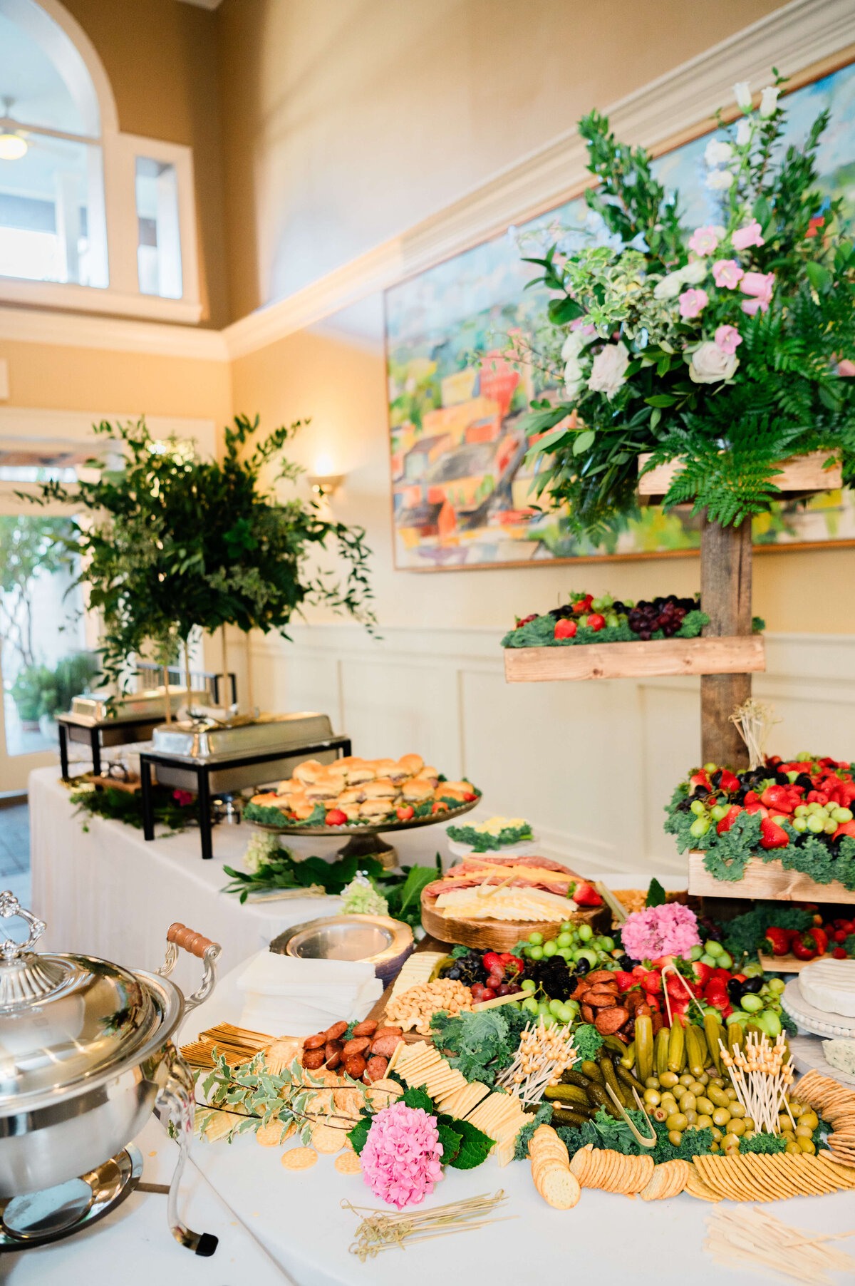 A buffet table with assorted cheeses, crackers, fruits, vegetables, pastries, and meats, decorated with fresh greenery and pink flowers, set in a bright room with a large floral arrangement and artwork on the wall.