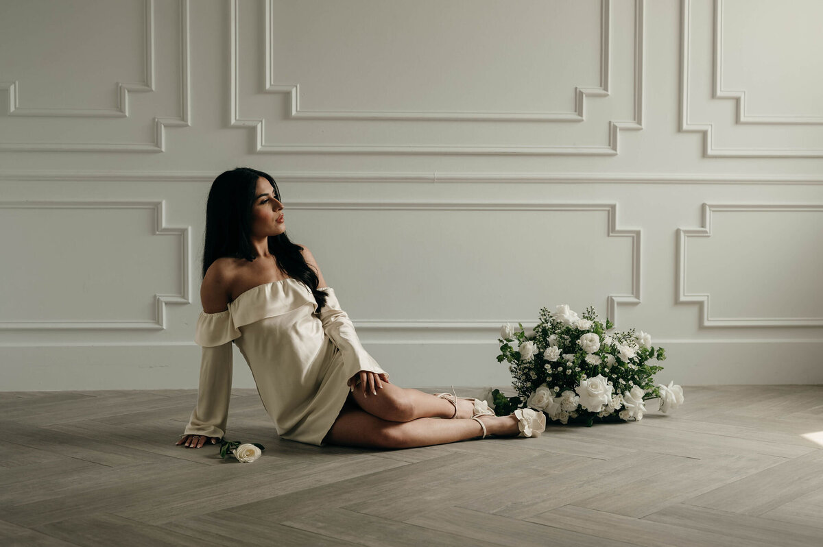 A woman in an off-shoulder cream dress sits on a floor by a white paneled wall. She gazes thoughtfully, with white flowers beside her and one in hand.