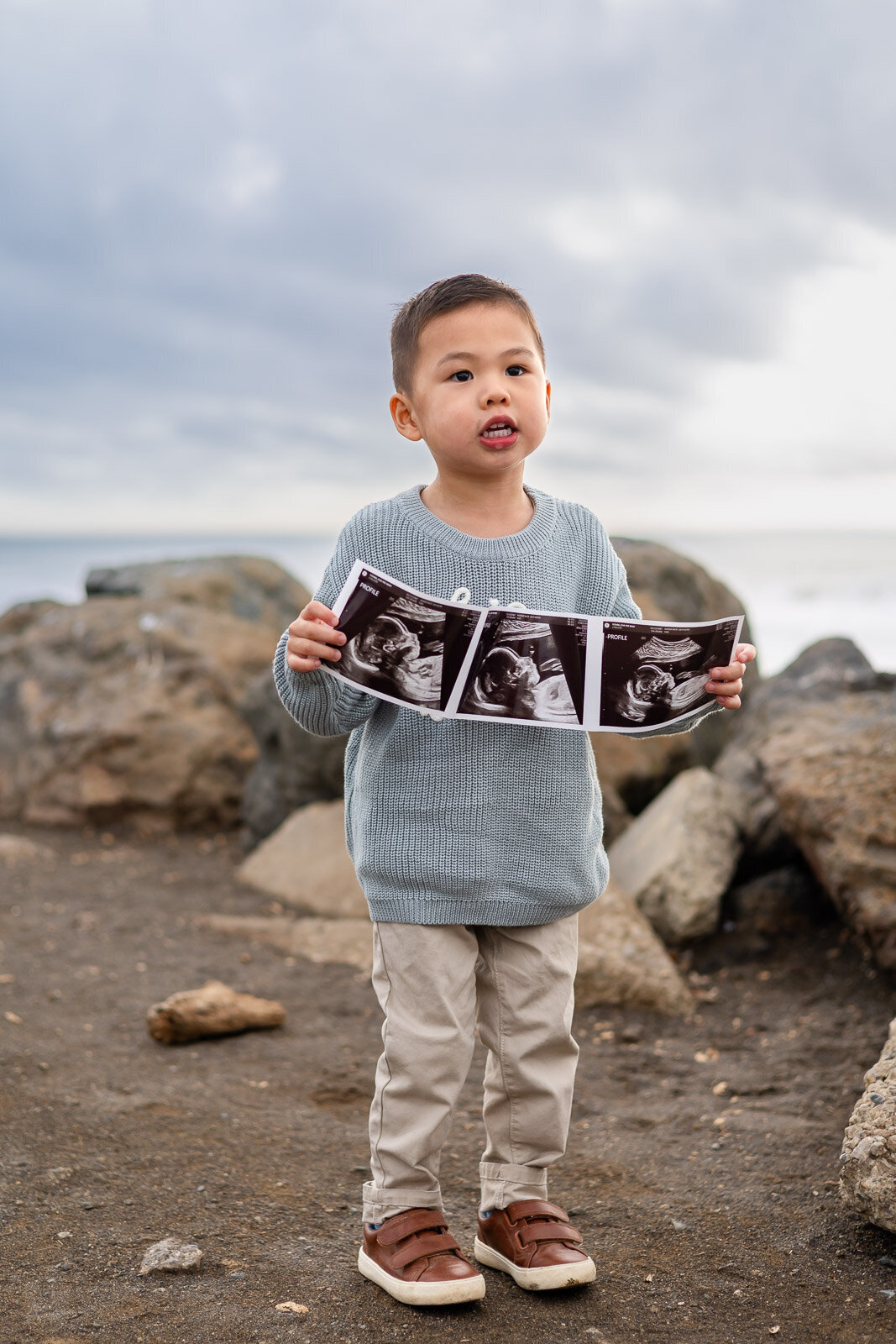 Toddler holding baby ultrasound photos at a rocky San Francisco beach, part of a Bay Area Maternity Portfolio – Ellobelle Photography