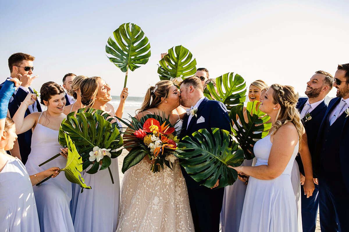 A bride and groom kiss while the wedding party hold up their monstera leaf bouquets