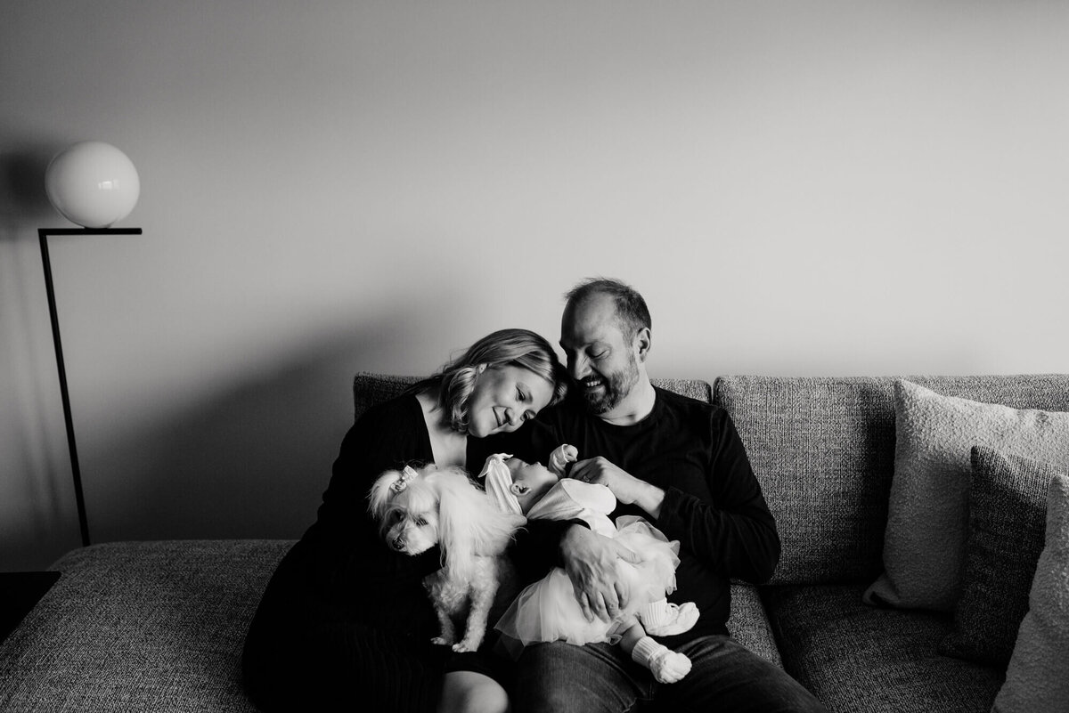 A black and white image of a family sitting on their couch with their little white dog and new baby girl, the mother and father look lovingly at their daughter during their photoshoot with Boston Newborn Photographer, Allison Wolf