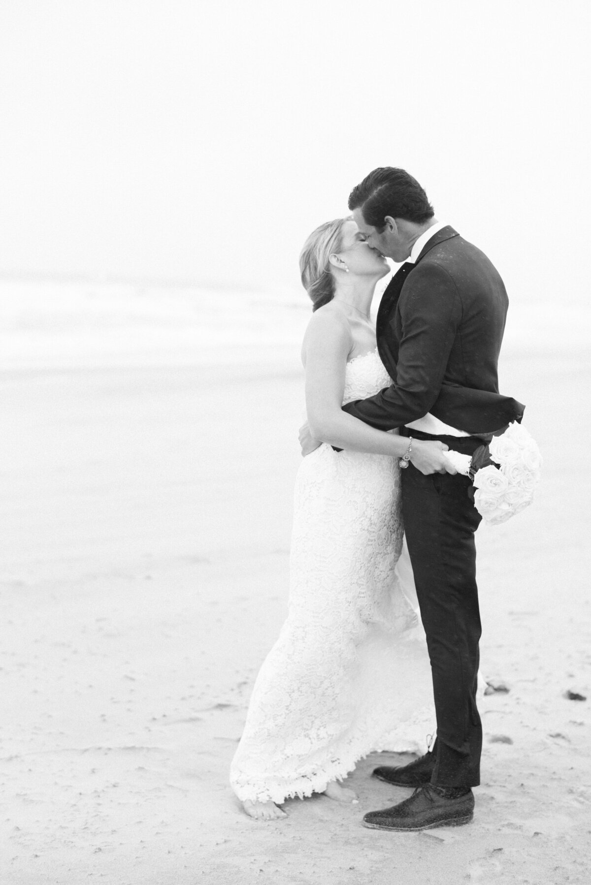 A black-and-white image of the newlyweds sharing a romantic kiss on the sandy shore. The bride, in a lace gown, holds a bouquet of white roses while the groom hold her in a dark suit.