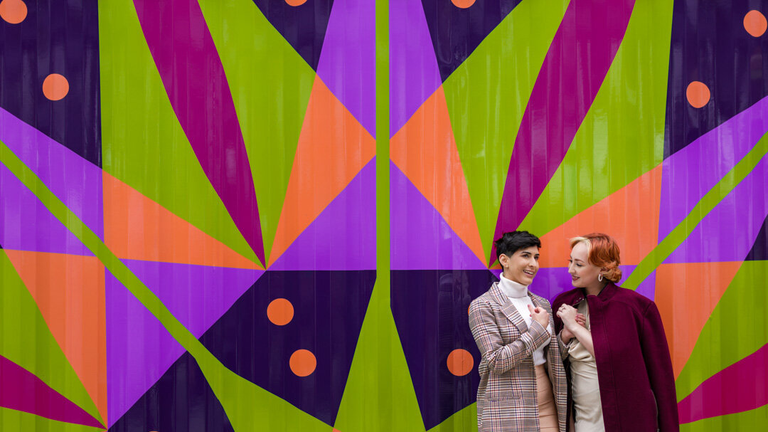 Same-sex engaged couple standing in front of a bold geometric graphic wall during their Saint Louis engagement session, showcasing their love and unique style in a vibrant urban setting.