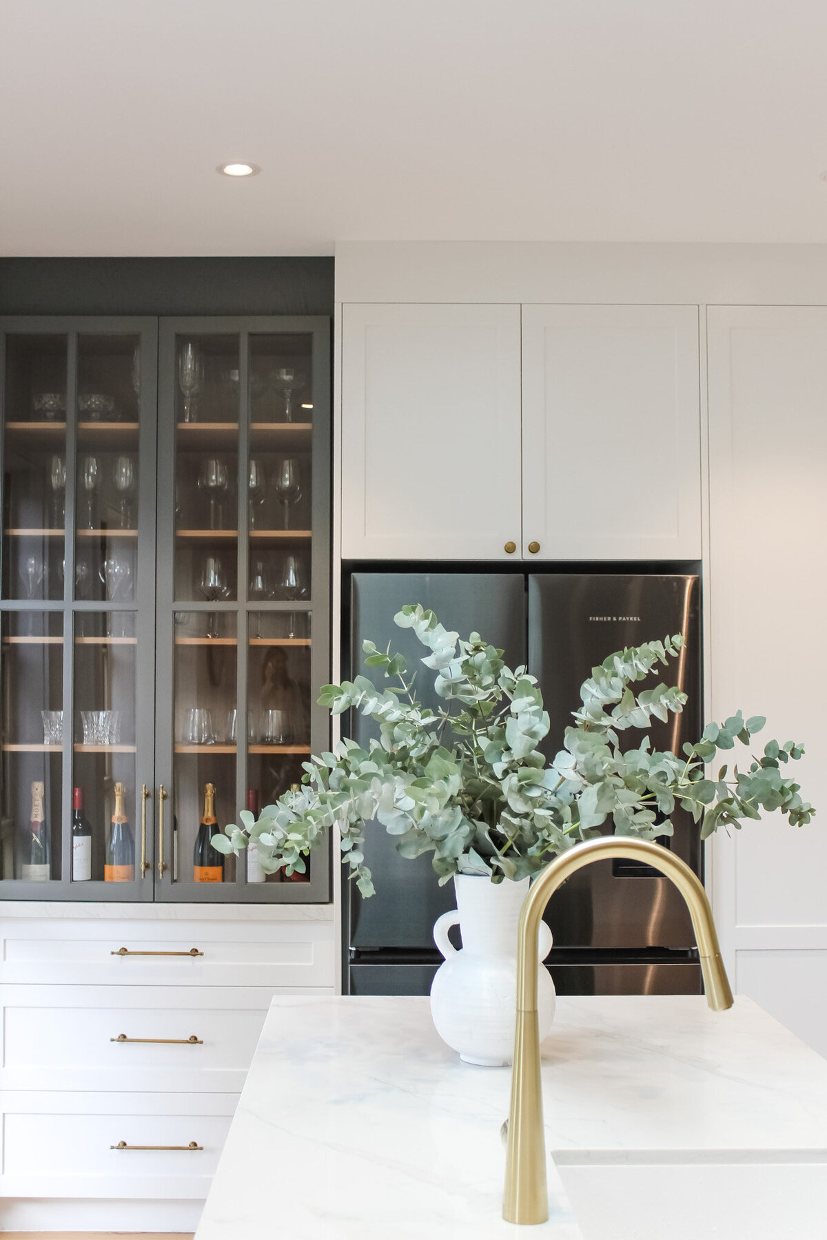 Modern kitchen with a marble island, gold faucet, glass cabinet filled with bottles, and a vase of greenery, masterfully crafted by a bespoke interior design team in Newcastle NSW.