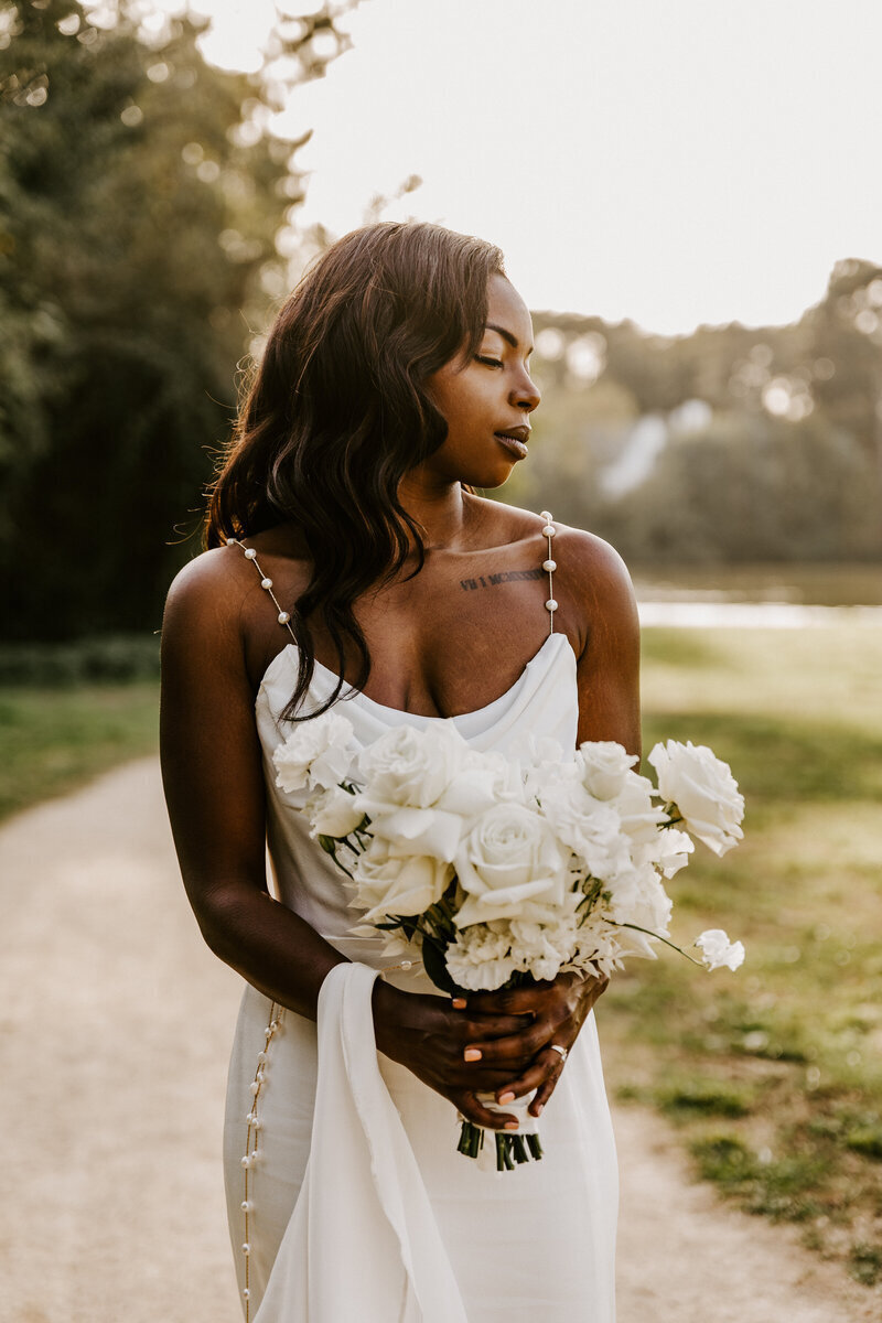 Mariée posant de face pour un shooting photo mariage, avec son bouquet de fleurs blanches, les yeux fermés dans un décor de verdure.