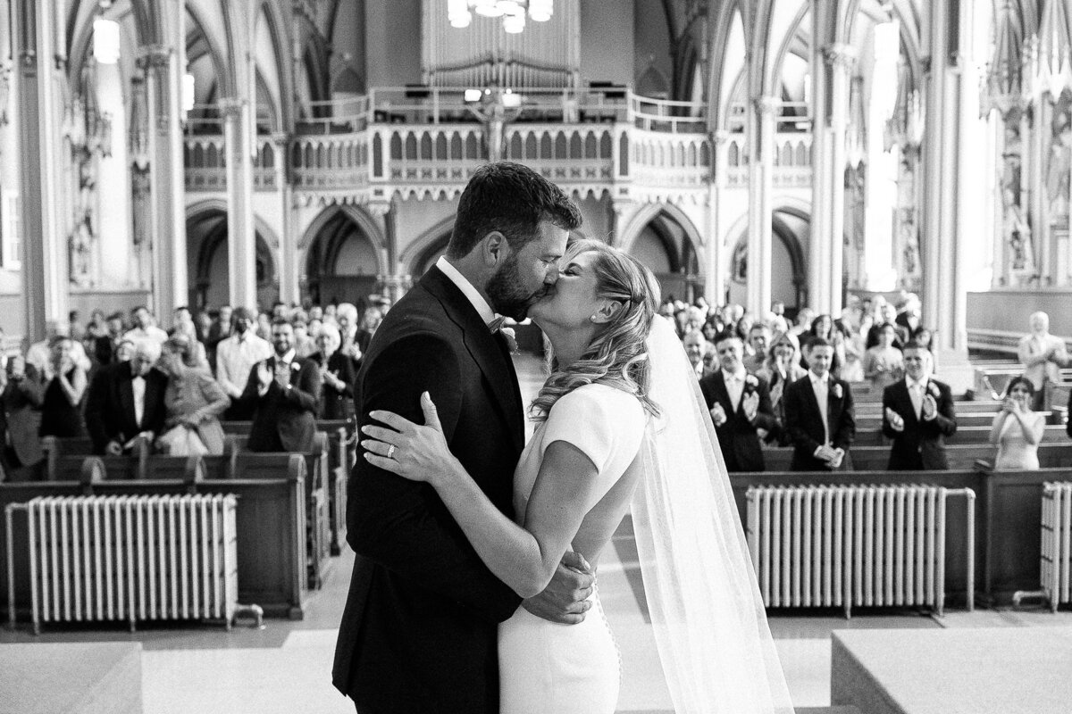 Baiser des mariés à l'hôtel de l'église avec leurs invités qui les félicitent en arrière. 