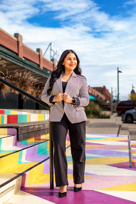 A young woman standing outside The Terminal in Pittsburgh wearing a plaid or checkered blazer and dark pants. She's positioned near colorful painted steps or a rainbow-colored surface, with buildings visible in the background.