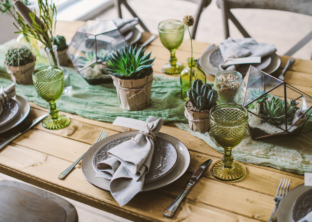 Rustic dinner set up on a wooden table with cactus, green goblet glassware for a party or event.