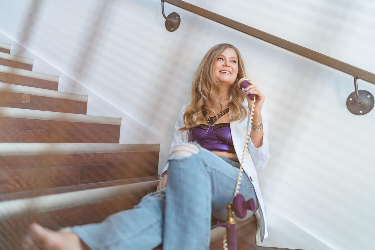 Female coach sitting on staircase holding phone to her ear, wearing jeans and purple top.
