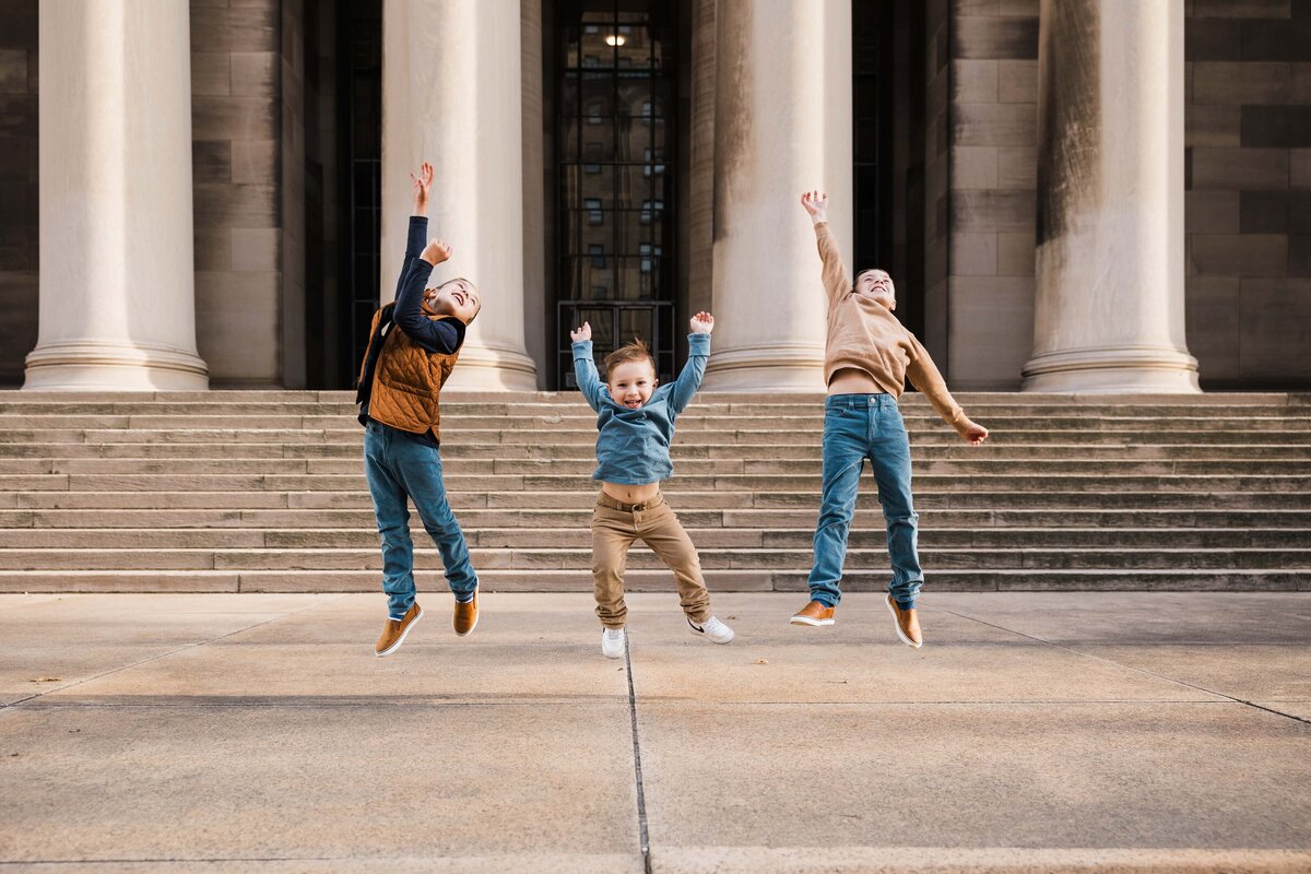 Three children joyfully jumping in front of a building with large columns, captured by a Pittsburgh family photographer.