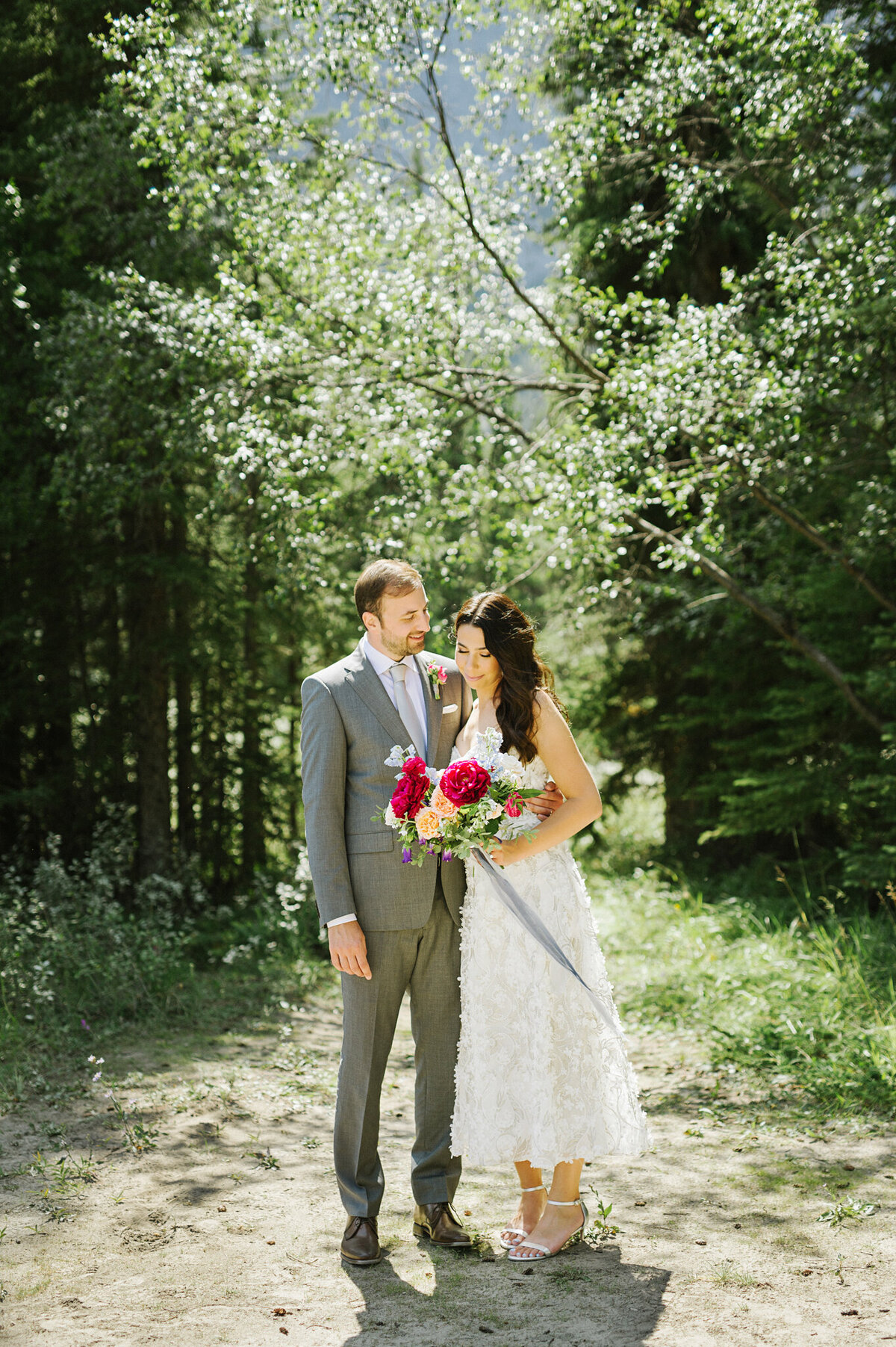Wedding couple standing together in Calgary park, bride wearing a trendy mid-length lace gown holding a fuchsia and peach floral bouquet, groom wearing a classic grey suit and pink boutonniere, captured by Christy D. Swanberg Photography, editorial elopement and wedding photographer in Calgary, Alberta, featured on the Bronte Bride Vendor Guide.