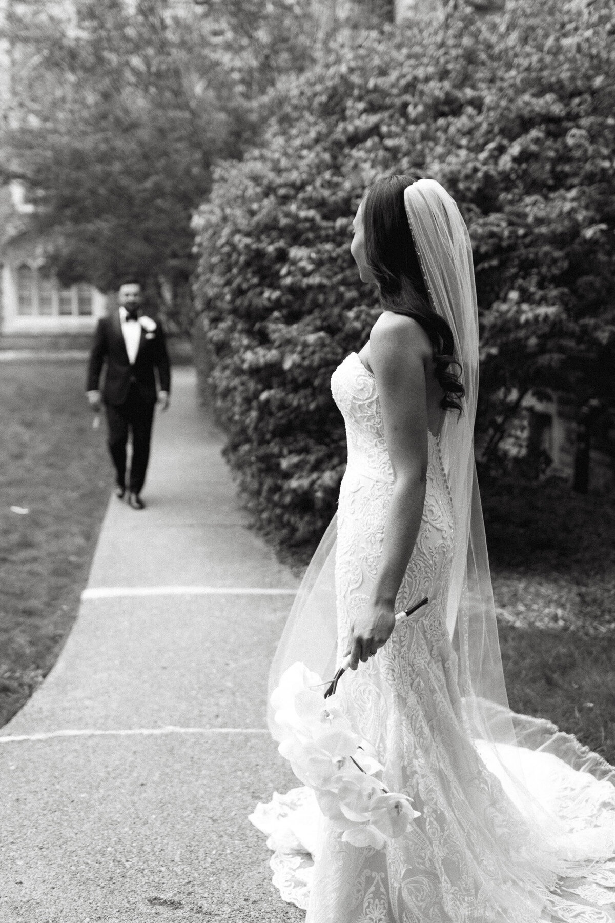 In this black and white image, a bride in a white lace gown and veil elegantly walks toward a man in a suit along a path lined with lush bushes and trees, captured expertly by a Toronto destination wedding photographer at Knox College.
