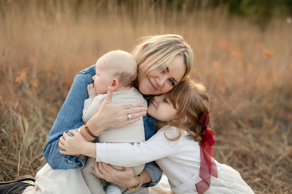 a mother holds her new baby into her chest while gently smiling as her daughter gives her a big hug