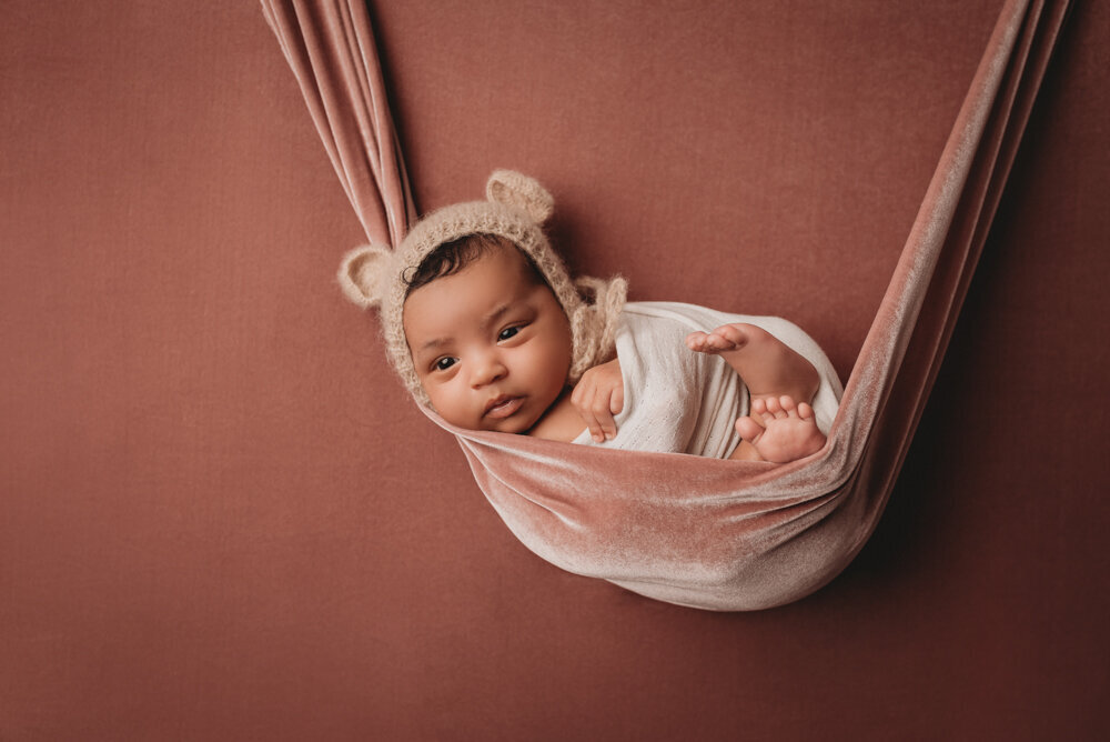 Atlanta, Georgia newborn photographer photographing Newborn baby girl swaddled in white wrap with knitted bear hat on in mauve swing and mauve background