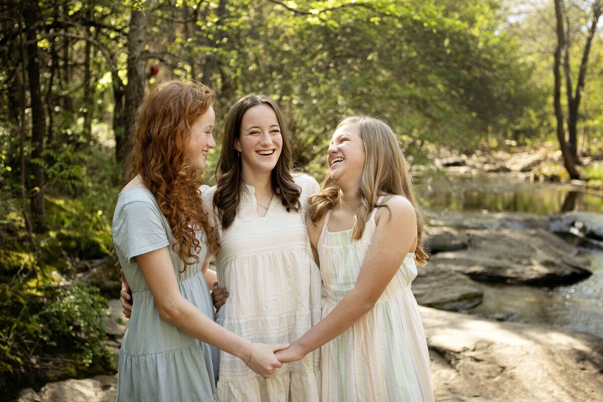 The three sisters embracing each other by the creek in Flower Mound, posing for a photo with the beautiful landscape behind them