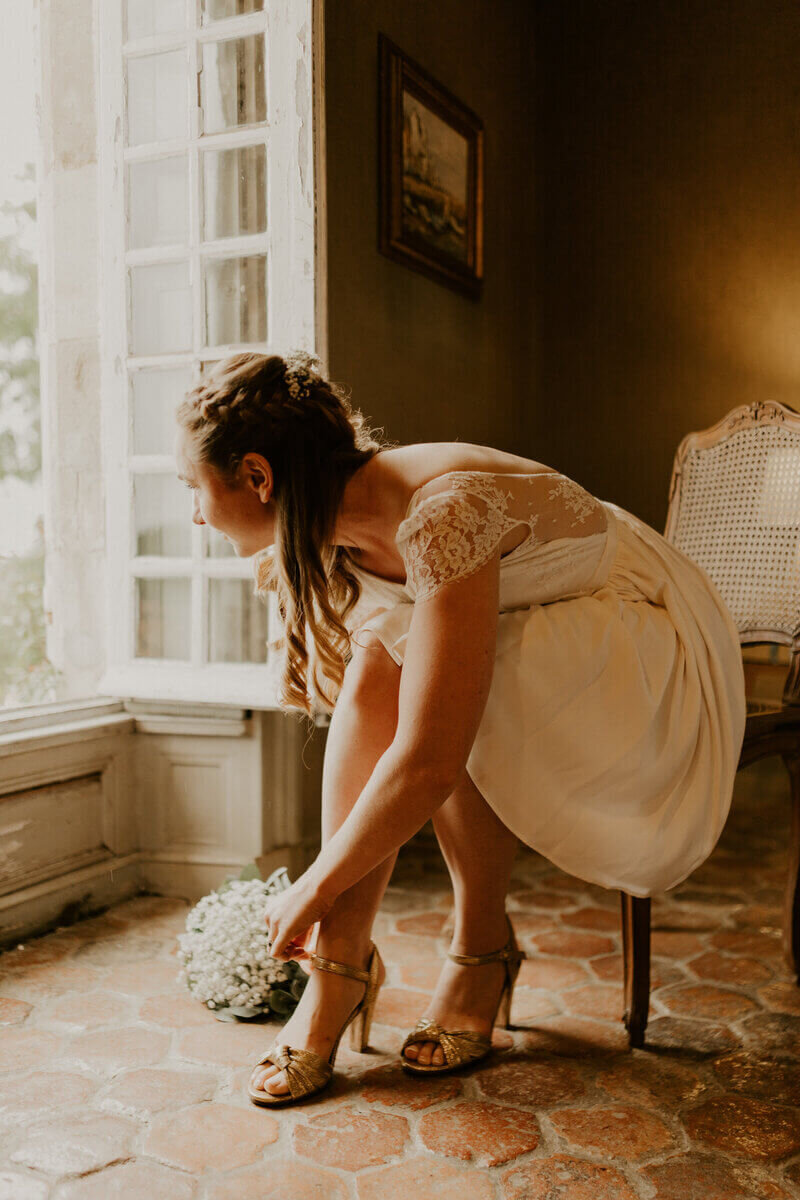 Mariée assise sur une chaise en bois reposant sur un sol en tommette, ajustant la sangle de sa chaussure et regardant par la fenetre ouverte lors d'un shooting photo mariage en Vendée.