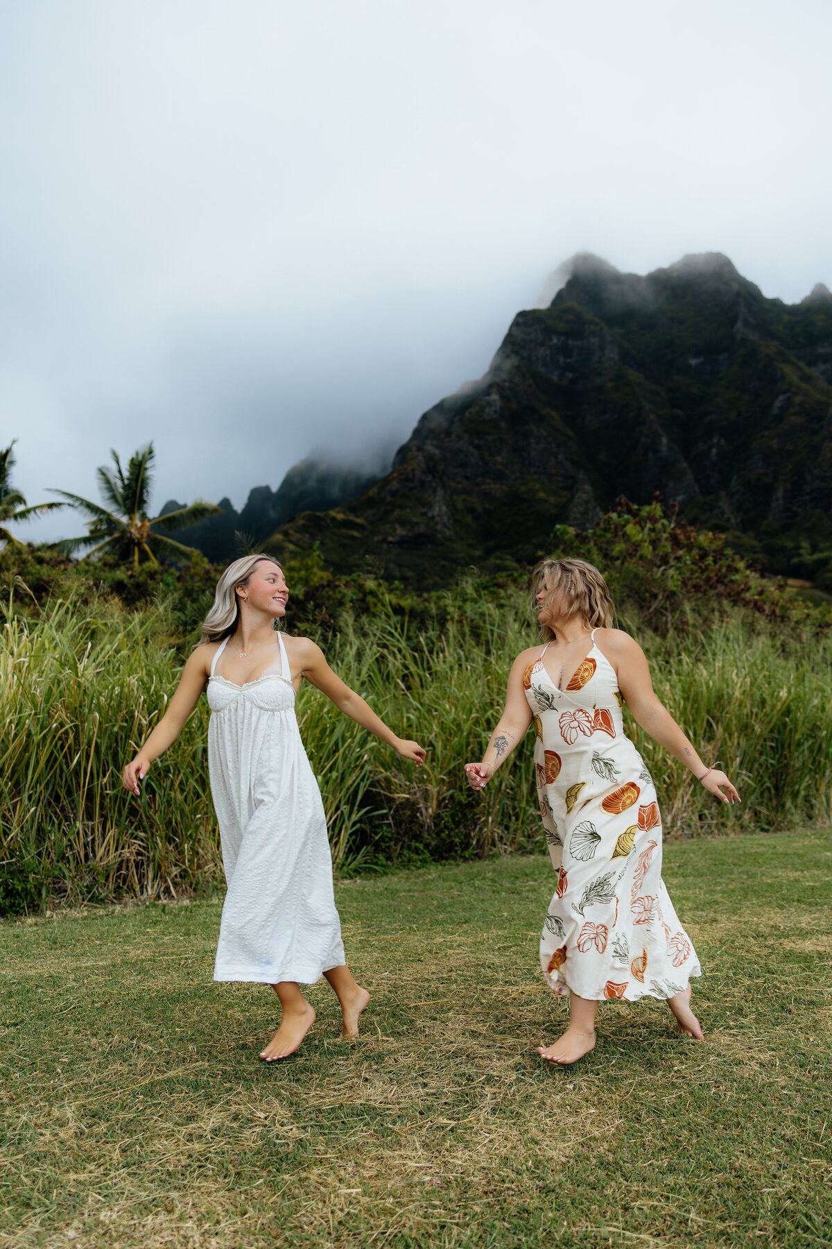 Two best friends twirling and holding hands in front of the lush green mountains of Oahu during a cloudy day photoshoot.