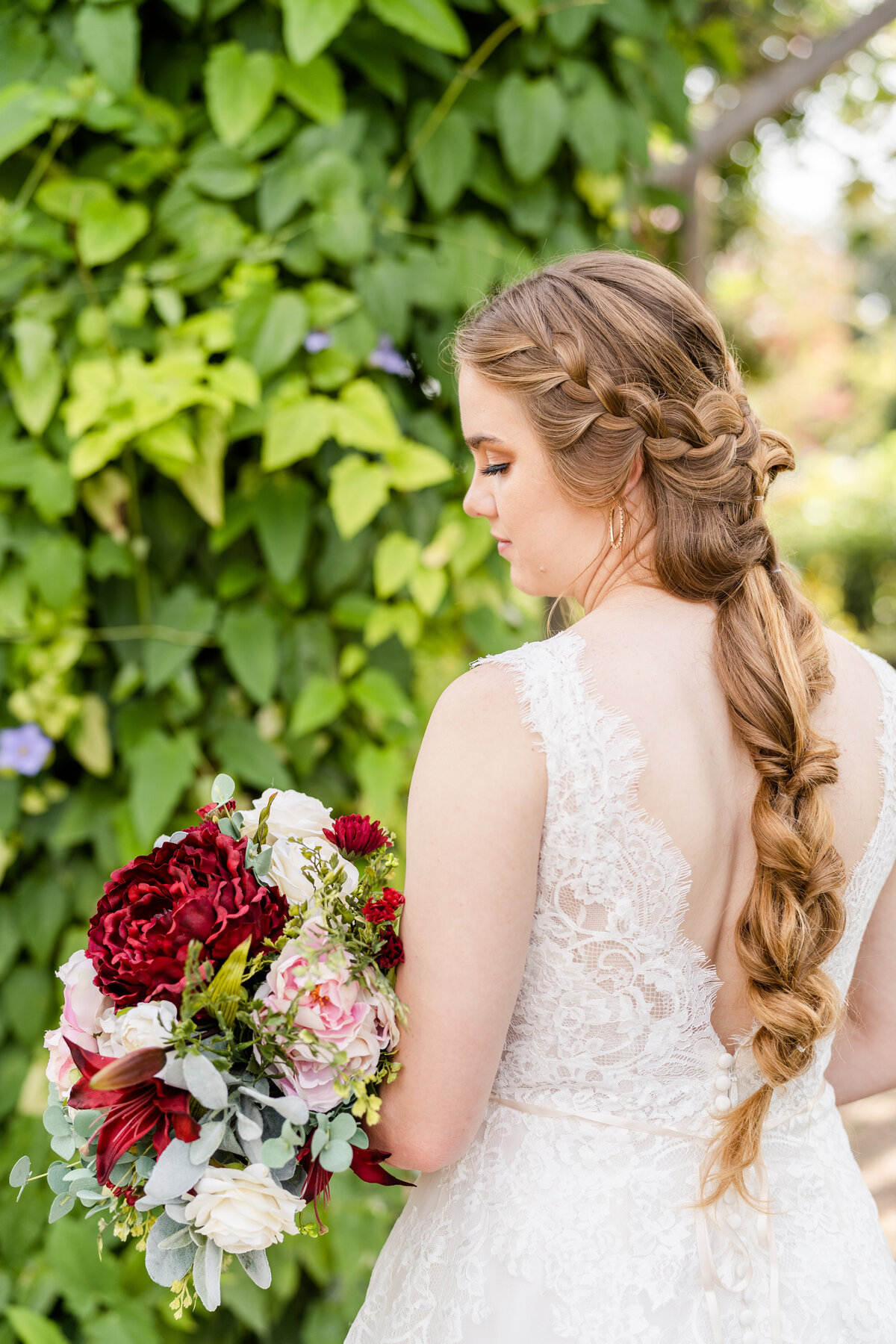 Bride with braided hair looking at bouquet  surrounded by greenery