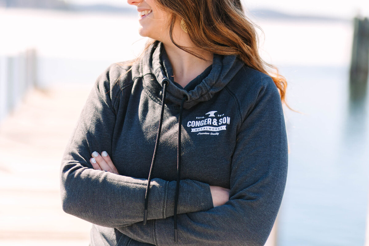 A woman wearing a dark gray hoodie with "Conger & Son Metalworks" printed on it stands with her arms crossed near a waterfront. Sunlight highlights her wavy brown hair as she smiles, with a blurred dock and water in the background.
