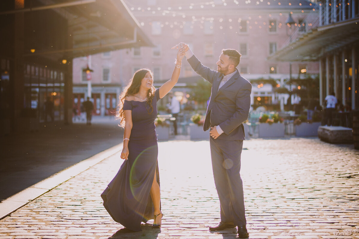 A couple twirling during their engagement session wearing formal clothing on a cobblestone street in the seaport district in nyc