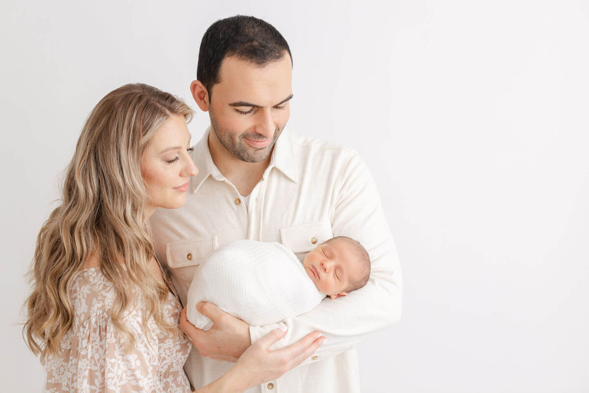 Mom and dad dressed in light, neutral tones are holding their baby and smiling down at him during their baby's newborn photo session in PDX.