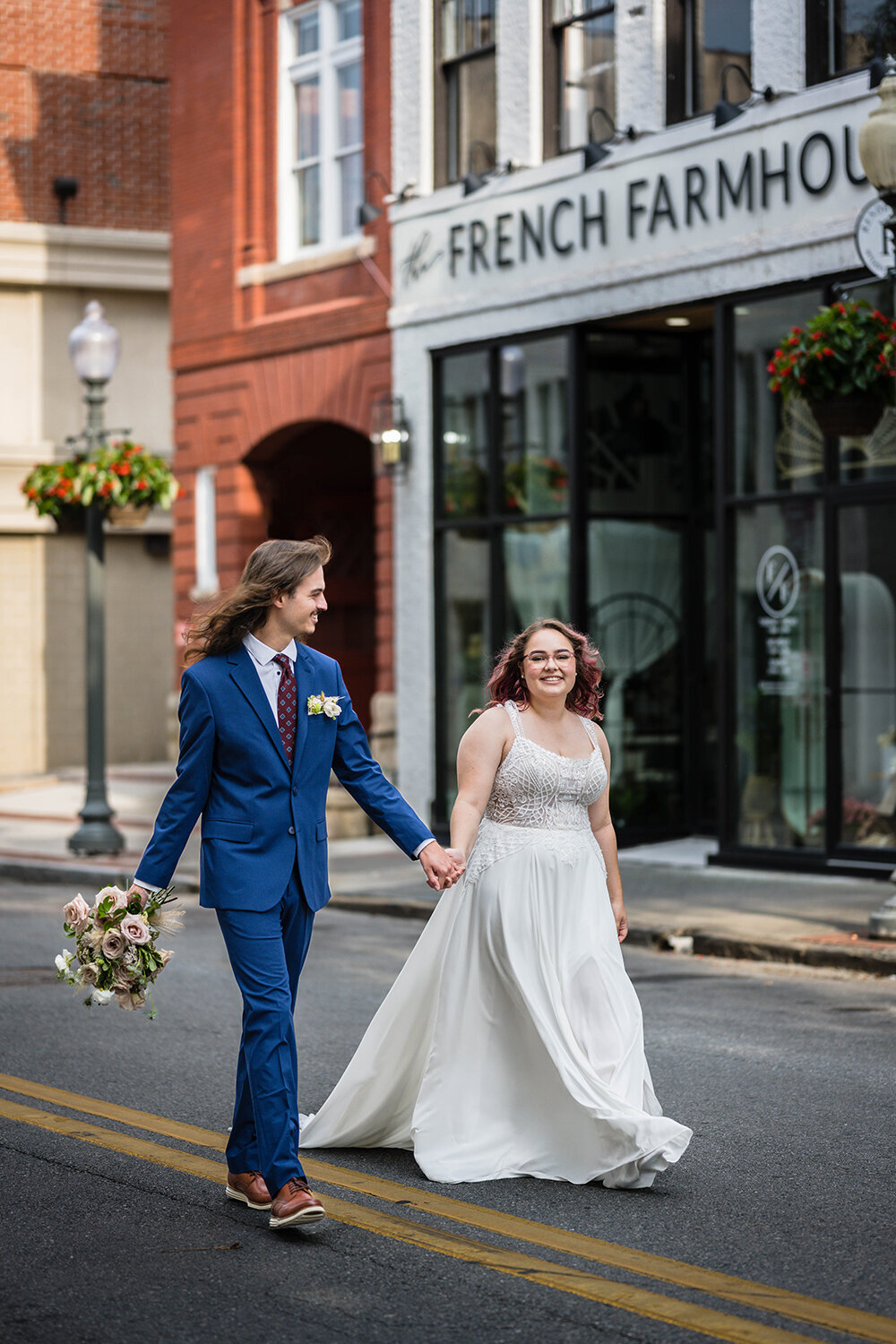 A newlywed couple walk hand-in-hand in the streets of Downtown Roanoke on their wedding day, with the Fire Station One hotel and the French Farmhouse store behind them. The bride walks holding onto her dress and smiling at the photographer while the groom holds the bride’s bouquet and smiles at the bride.