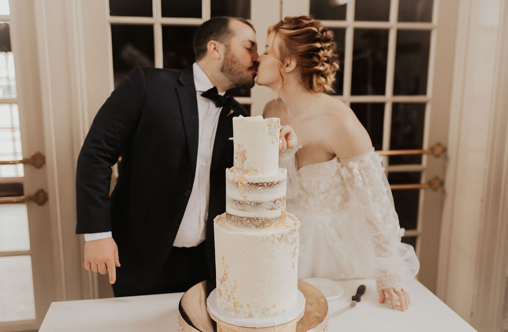 A couple in wedding attire share a kiss while cutting their wedding cake. The cake sits in front of them with white frosting, pastel accents, and gold leaf.