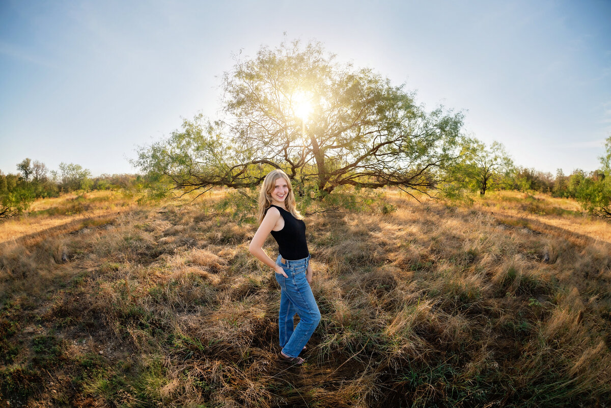 A high school senior girl poses in front of a field with a dramatic tree behind her.