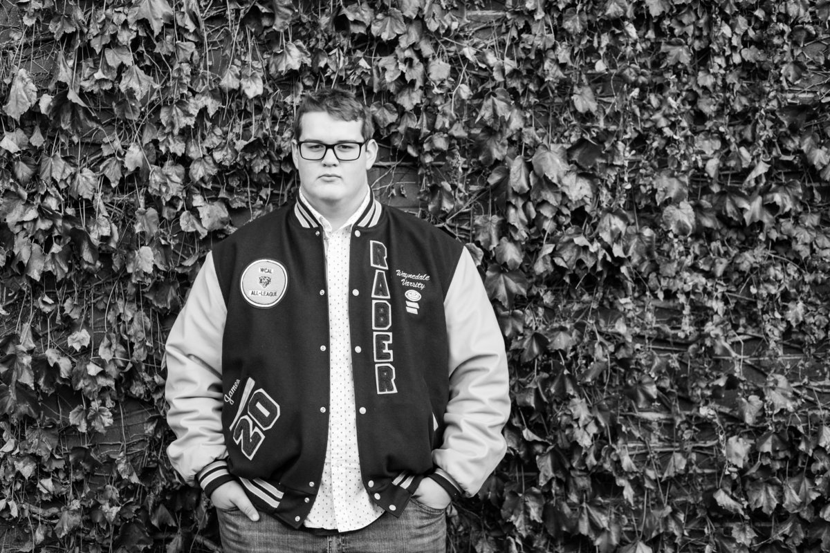 black and white photo of senior guy leaning up against a wall full of vines, he is wearing his lettermans jacket, photographed downtown Wooster, photographed by Jamie Lynette Photography