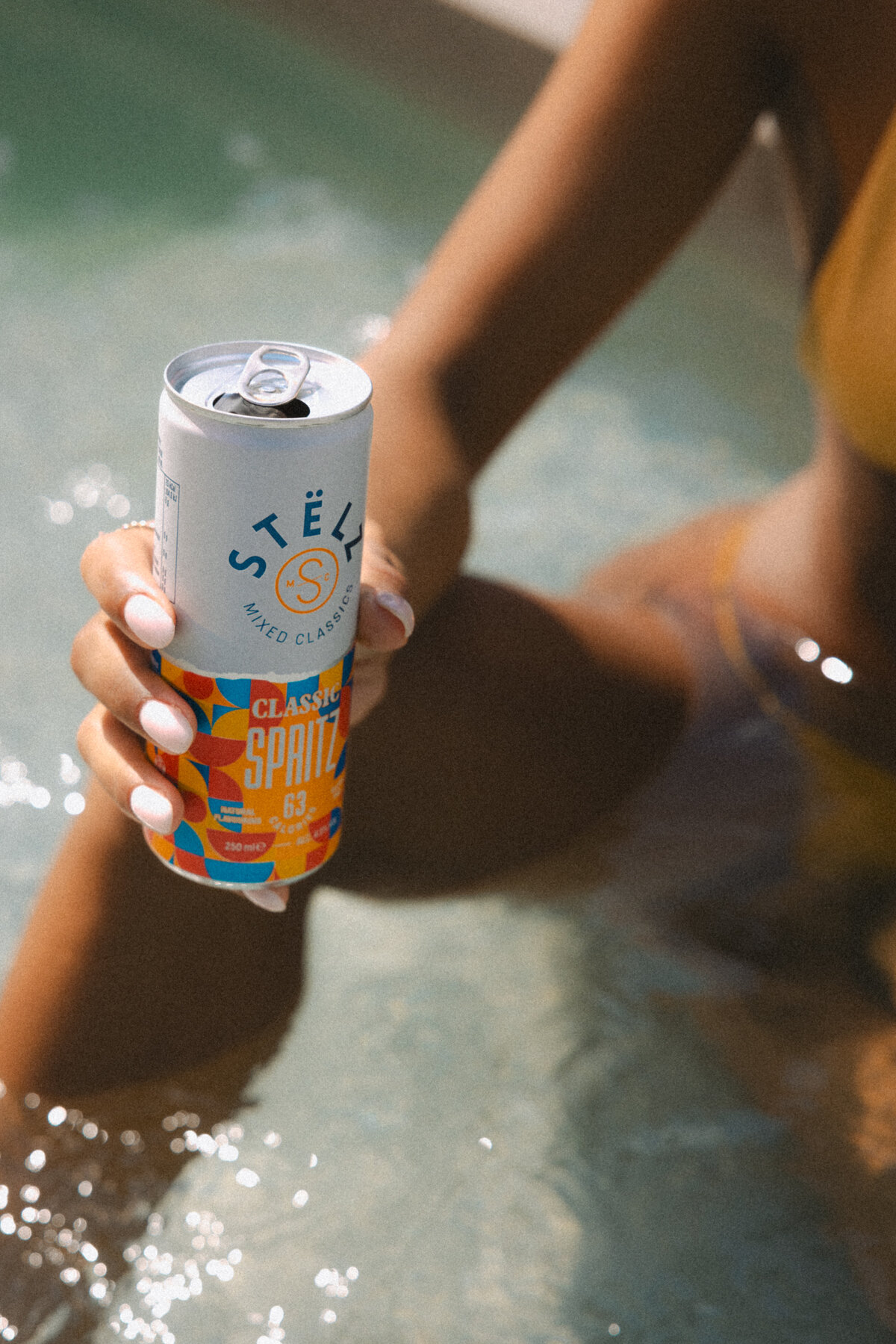 A woman sitting by the pool, holding a STËLZ Classic Spritz can, as the sunlight casts a warm glow on her and the surrounding water. The scene exudes tranquility and the essence of summer. Photography by Bo Eppink.
