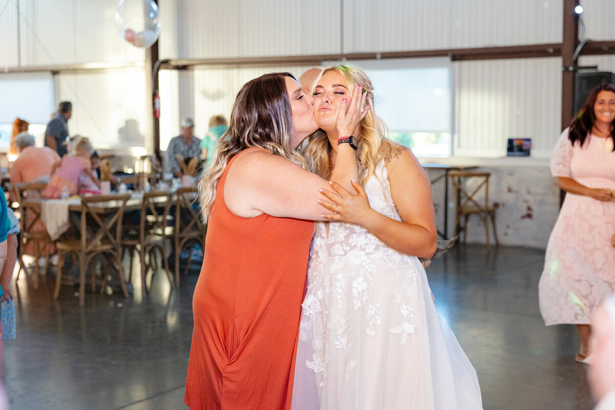 A guest playfully kisses the bride on the cheek while the bride makes a funny expression, enjoying the moment.