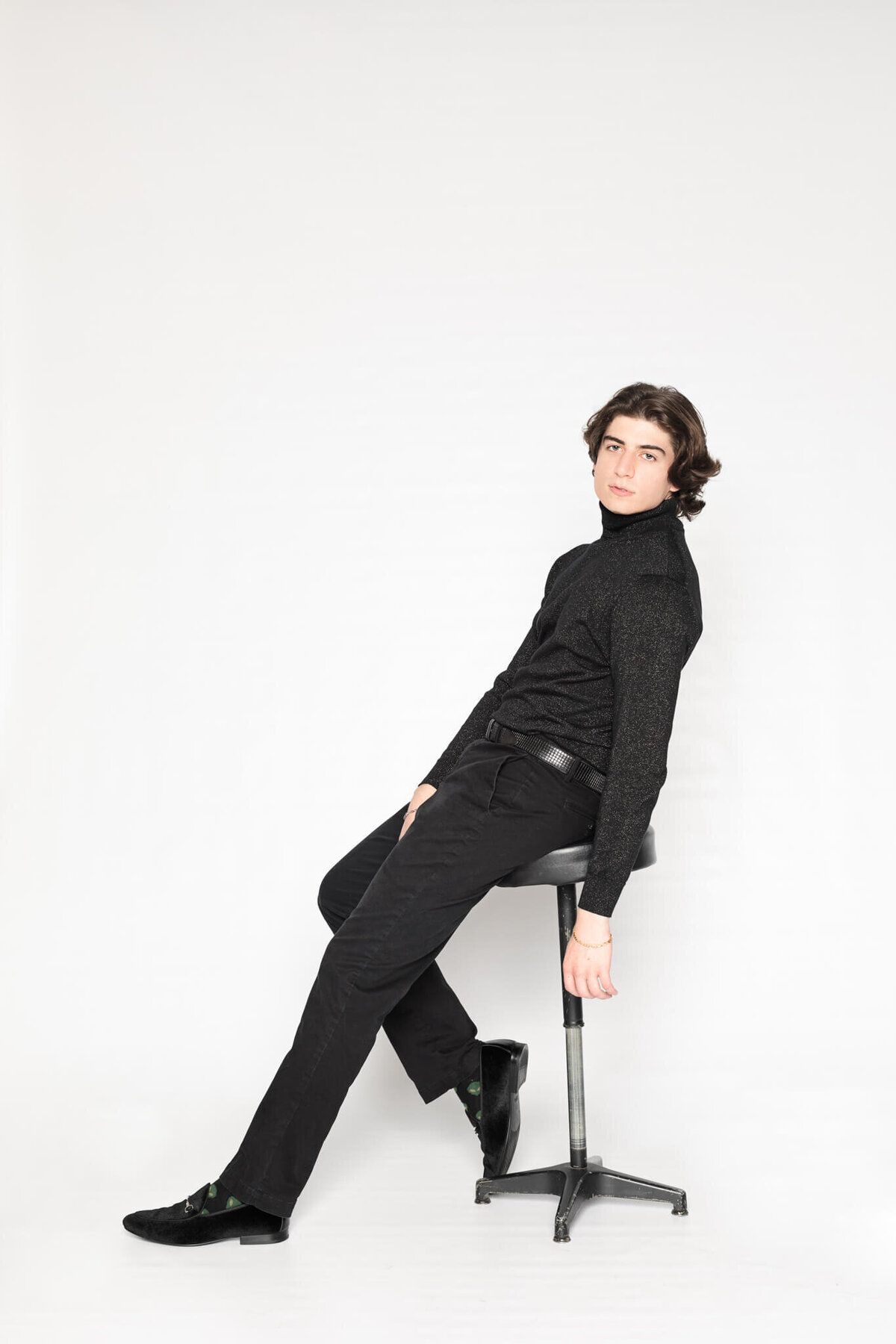 A person with shoulder-length hair sits on a stool against a white background, wearing a black turtleneck and black pants. They lean back slightly with one arm resting on their knee.
